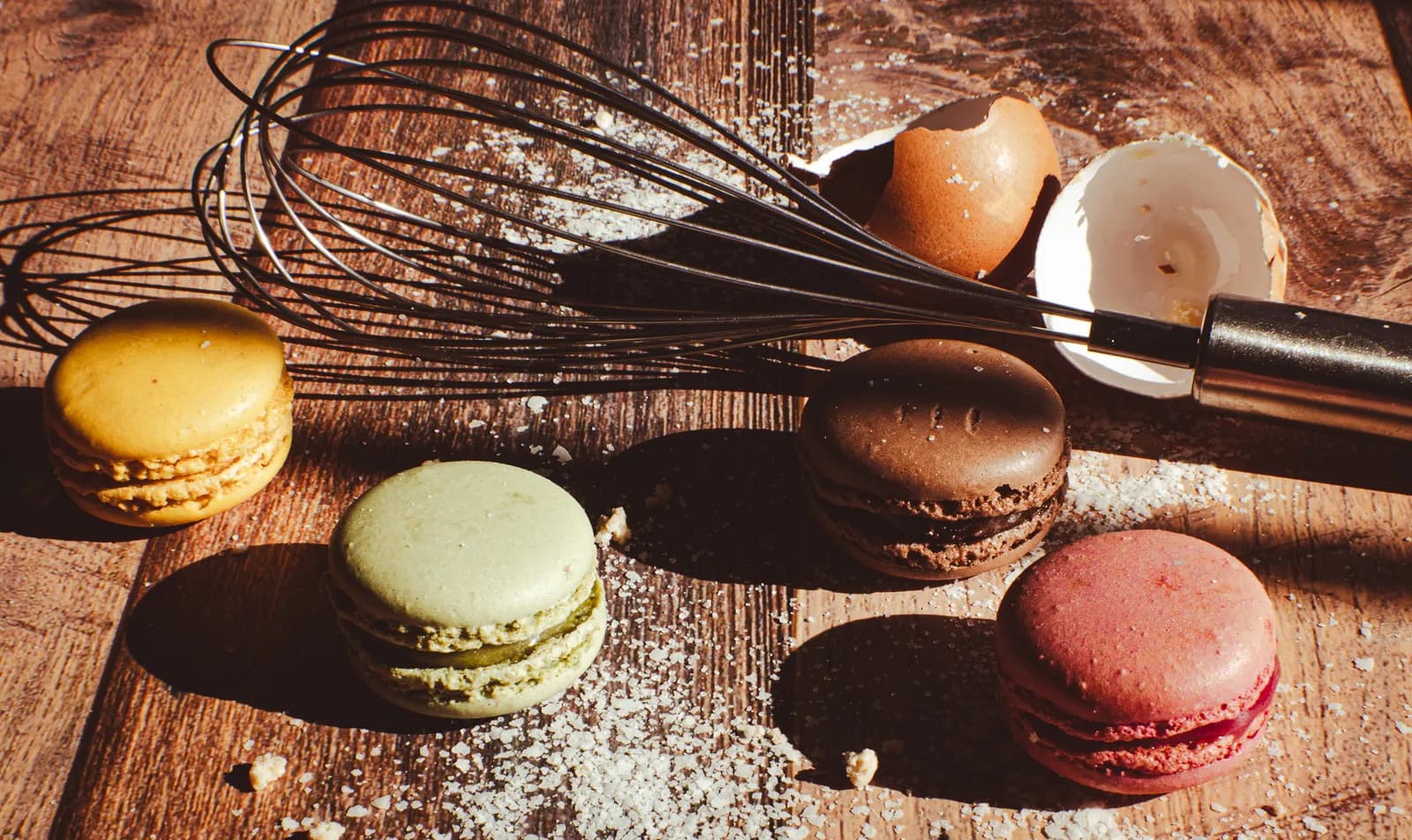 A wooden surface with an egg whisk, several macarons, a cracked eggshell, and sprinkled sugar or flour.